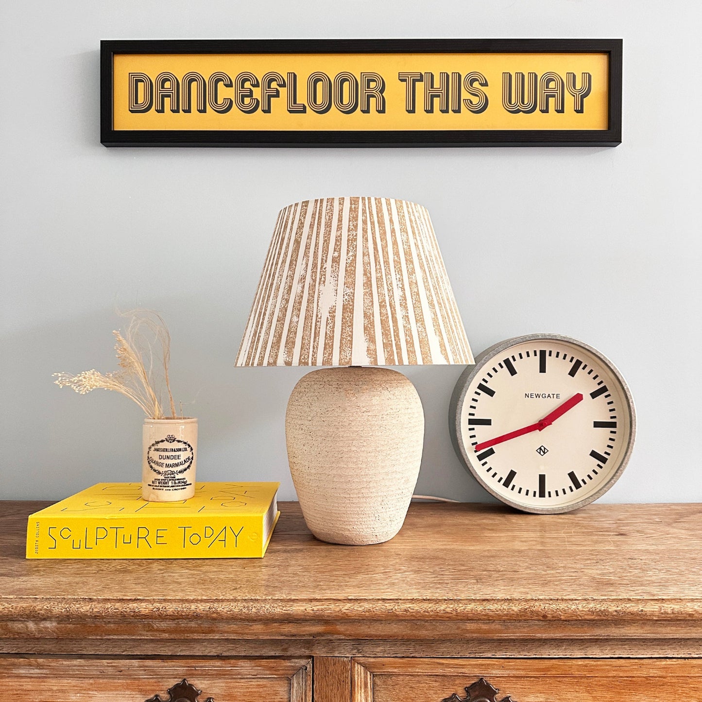 Bold stripe lampshade in warm neutral tones displayed on a rustic wooden sideboard, styled with a clock and books. A modern empire lampshade that combines striped lighting design with timeless elegance.