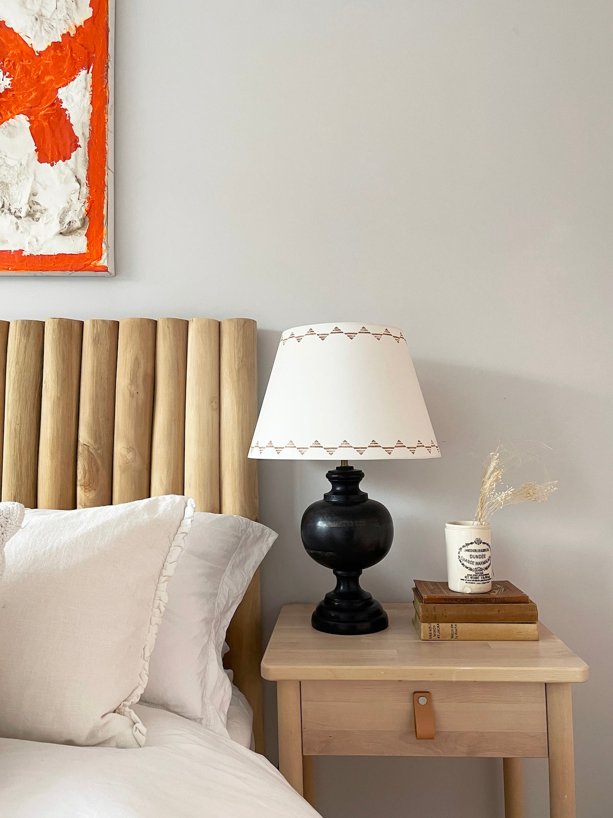 Empire lampshade for table lamps in cream with geometric triangle design, styled on a wooden bedside table with books, black lamp base and white bedding. There is bight orange and white artwork above the bed.
