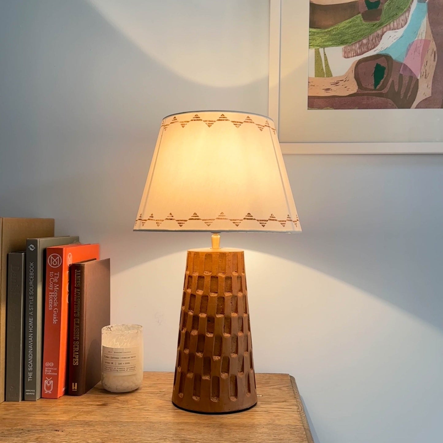 Bold empire lampshade with triangle pattern, glowing warmly on a wooden textured lamp base beside books and a candle for cosy lighting.