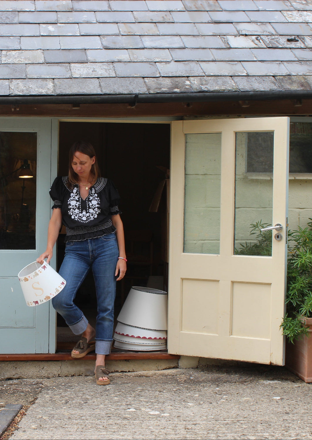 Grace Franks, founder of Studio Zef, standing outside her handmade lampshade studio in the cotswolds holding a bold lampshade. The image shows the creative workspace where colourful lampshades, empire light shades, and unique designer lighting are crafted in the UK.