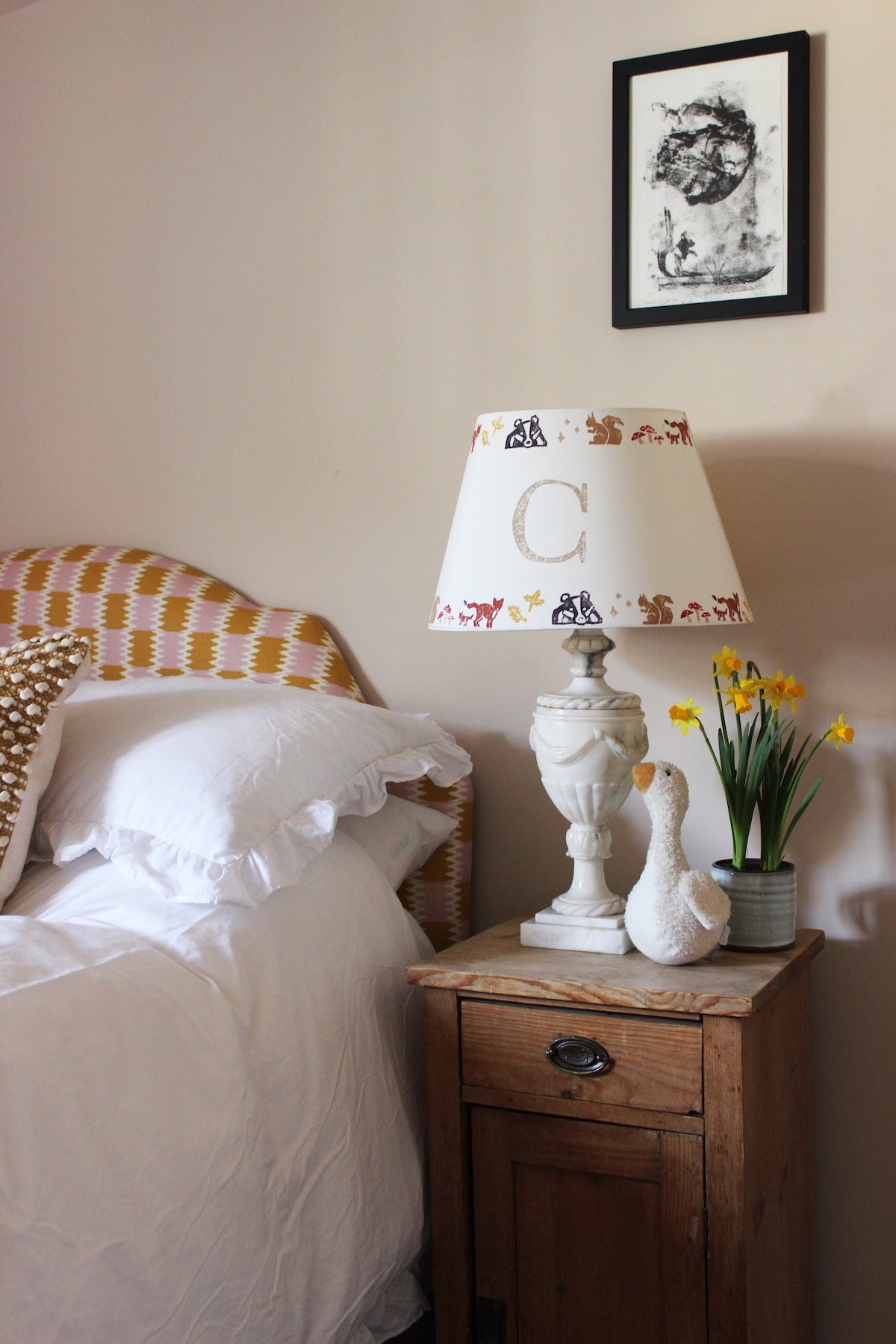 A cosy children's bedroom with a rustic wooden nightstand featuring a personalised lampshade with the letter "C" and a countryside pattern. A plush goose, daffodils, and white bedding add charm. Neutral empire light shade with pretty animal details, ideal for kids bedroom design and personalised kids gift.