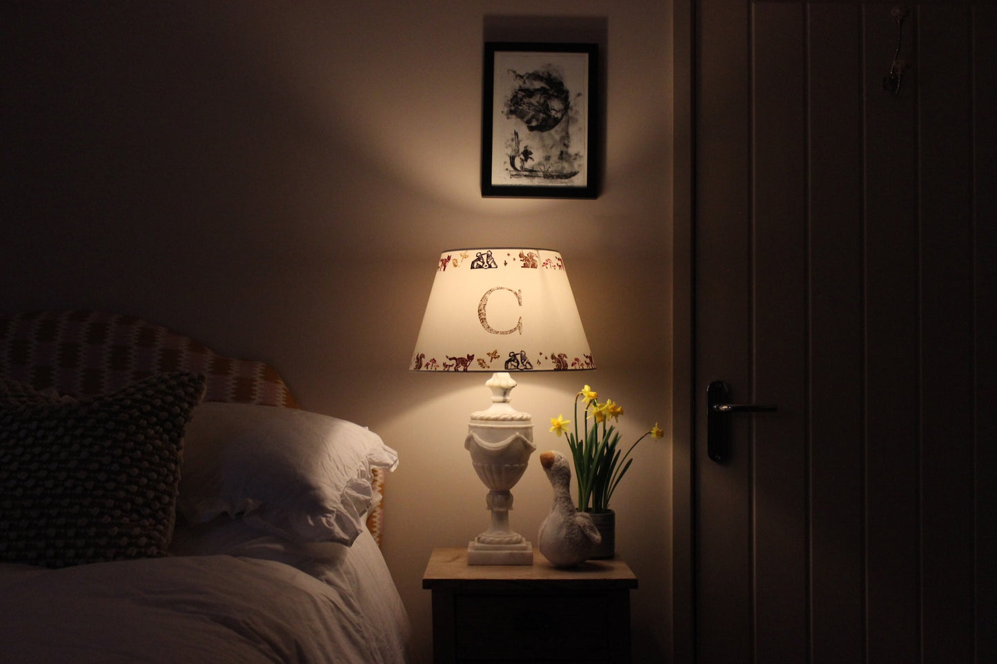 Personalised kids lampshade with woodland animals and letter C, glowing softly as part of cosy kids bedroom design, placed on a rustic wooden bedside table. The lamps is on giving the room a soft ,warm glow. A plush goose, daffodils, and white bedding add charm.