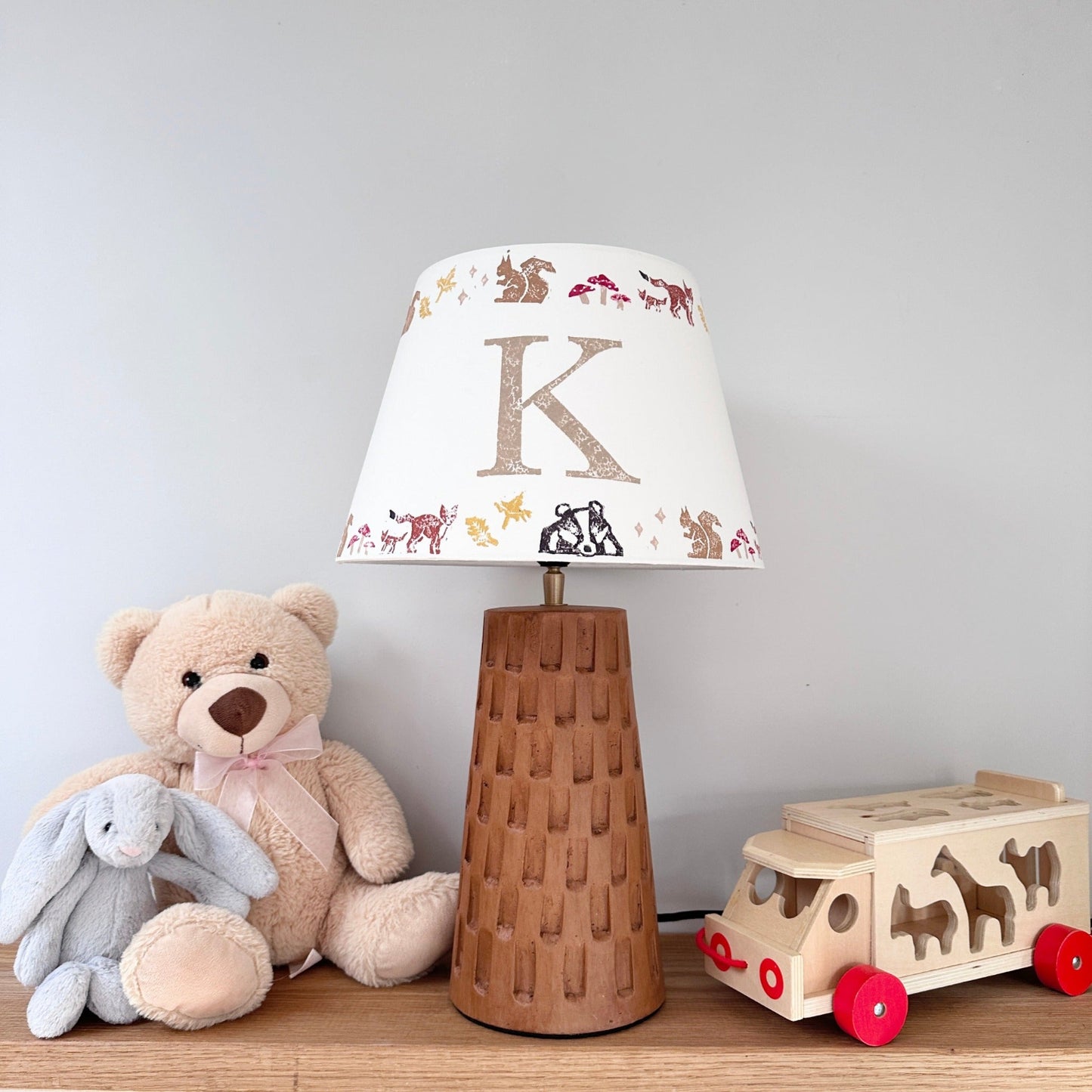 Personalised empire lampshade with woodland animals and letter K on wooden lamp base. Animal lampshade styled in kids bedroom decor with wooden truck and soft toys, ideal as a boys lampshade or kids lampshade. A personalised lampshade gift for new parents or christening present.