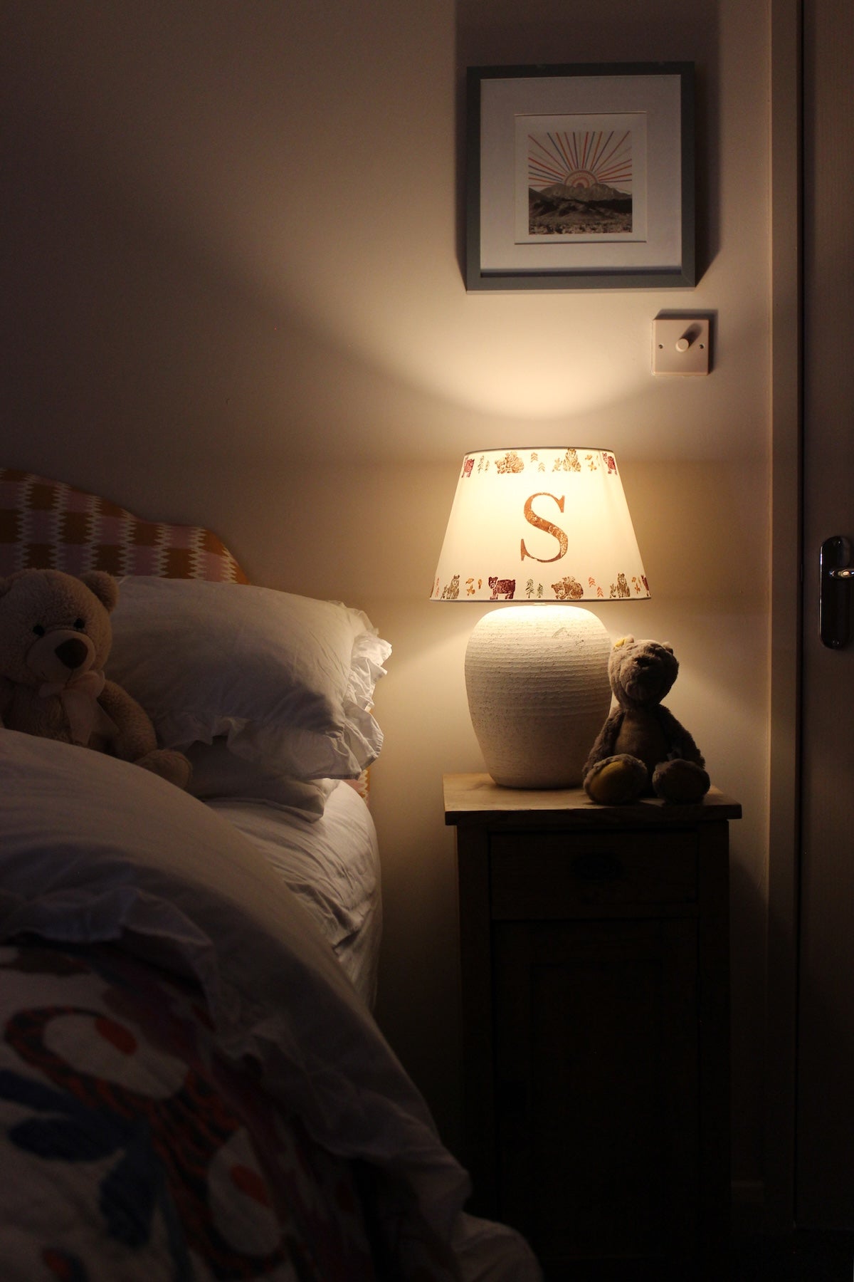Glowing personalised bear lampshade with letter 'S', styled as kids lighting on a bedside table with teddy bears. A warm and neutral empire lampshade for kids bedroom décor. The rustic bedside table adored with a ceramic lamp and lampshade. There are two teddy bears, one on the bedside table next to the lamp, the other folded into the duvet.