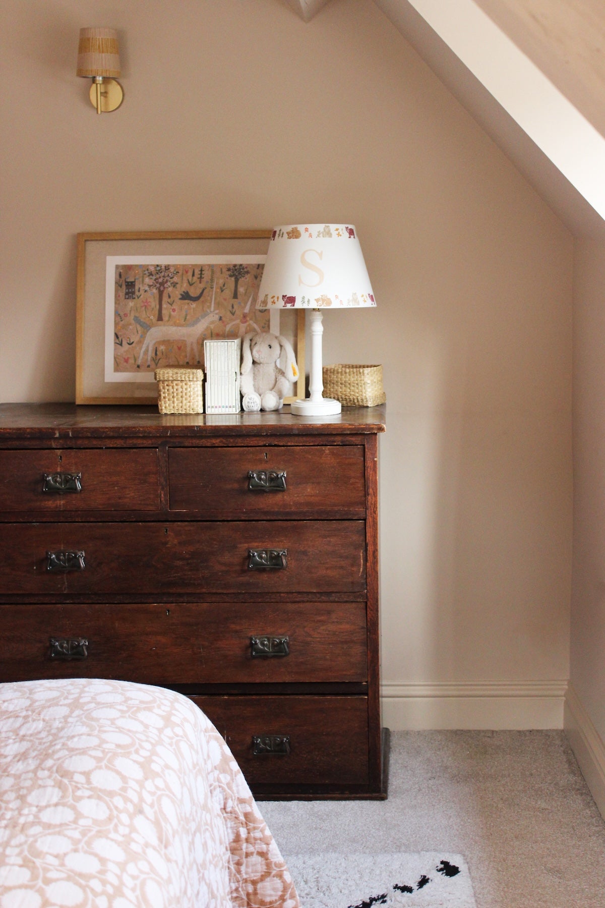 Cozy corner of a girls bedroom, featuring a wooden dresser and white table lamp paired with a personalised kids lampshade adorned with a bears pattern and the letter 'S'. A framed artwork hangs behind the lampshade featuring a unicorn and mythical setting.