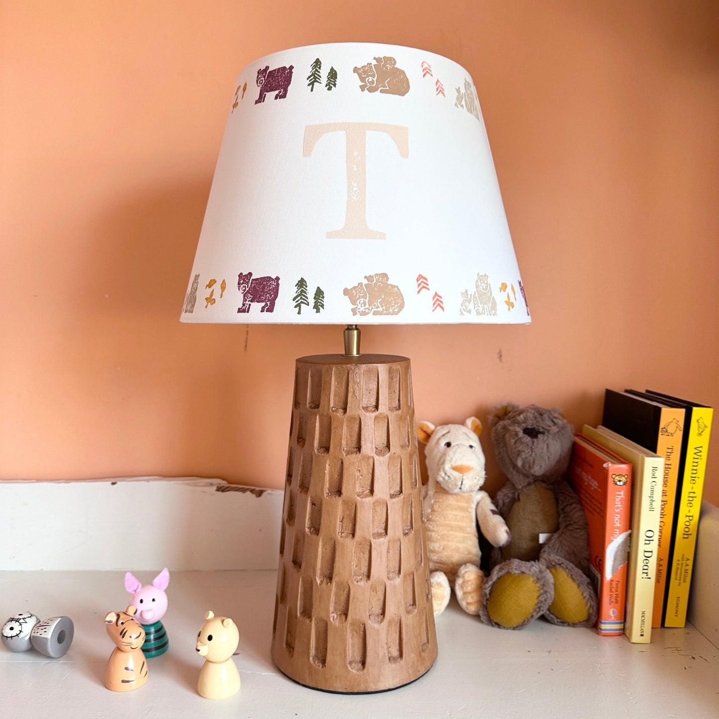 Personalised kids lampshade with grizzly bears and trees design, colourful empire lampshade with initial 'T' on a brown ceramic lamp base. A pretty lampshade for kids bedroom décor and new parent gifts. The lamp is in a kids bedroom chest of drawers next to stacked children's books, toys and soft toys.