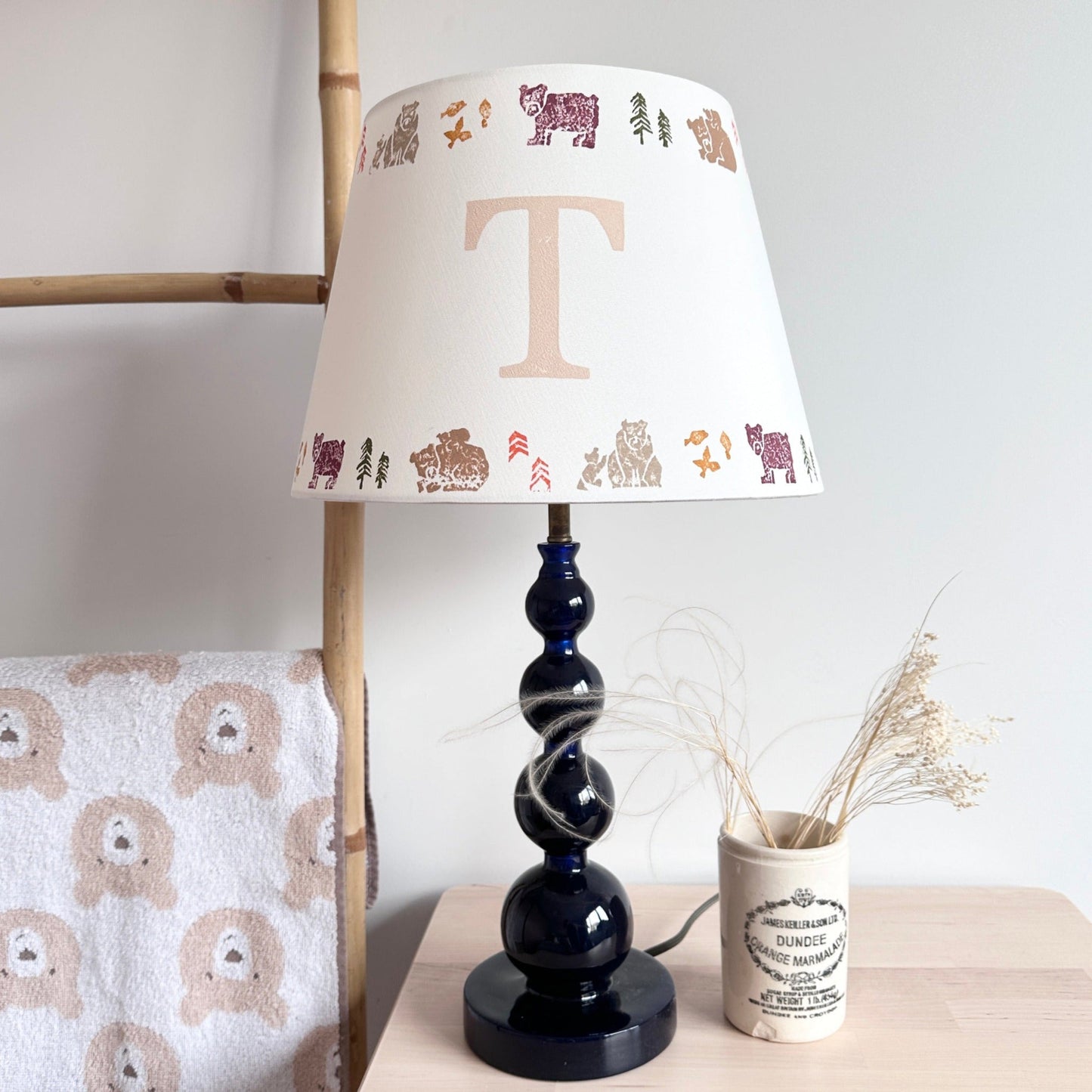 Empire light shade for kids bedroom with bear and woodland design, personalised with initial 'T'. A colourful lampshade and animal lampshade perfect for kids bedroom design and presents for new parents. The kids lampshade is on a blue bobbin lamp, placed on a wooden side table with antique ceramic vase and dried flowers.