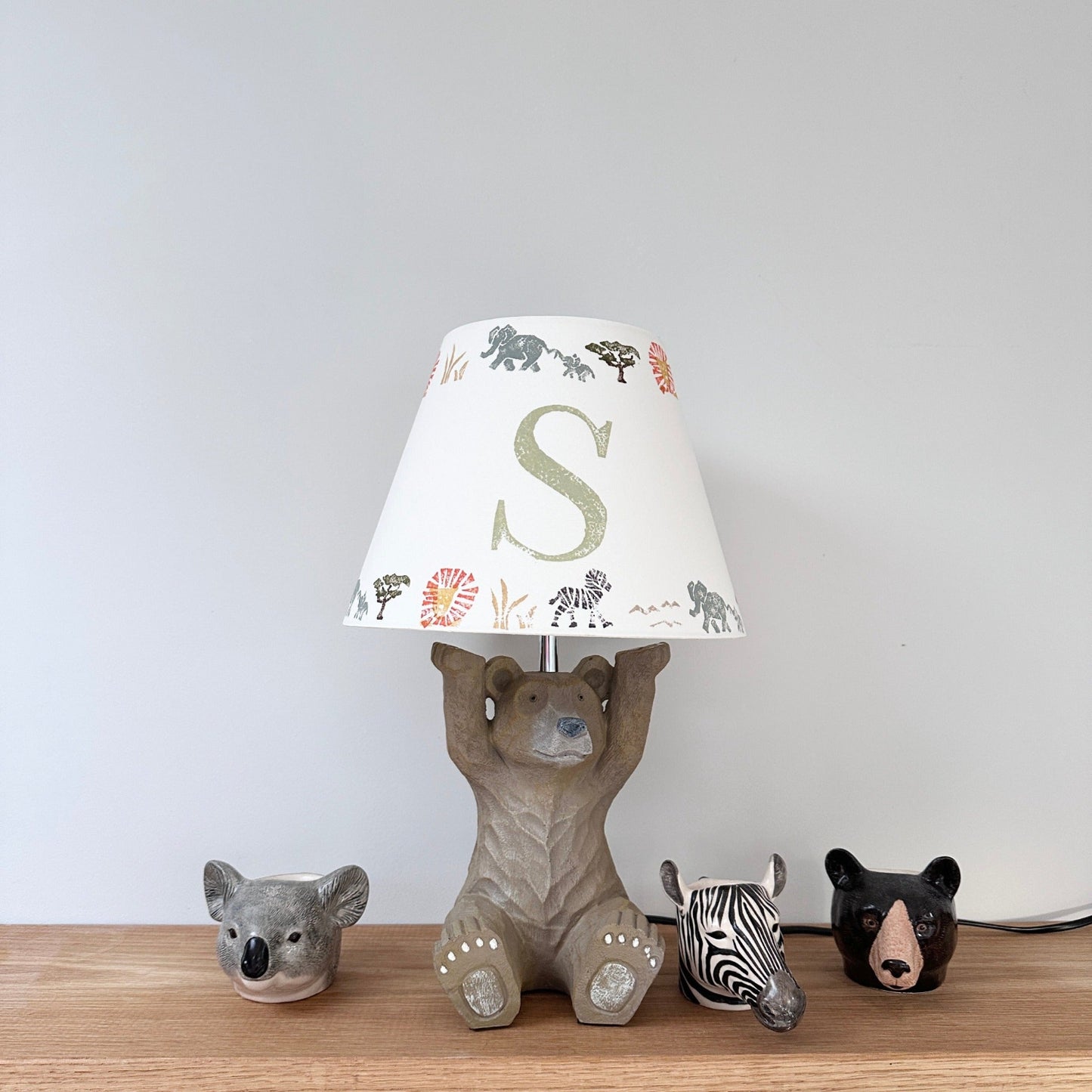 Personalised kids safari lampshade with letter S, colourful lampshade featuring elephants, zebras and lions. Empire lampshade design perfect for boys lampshade or kids lighting, ideal for kids bedroom decor, placed on a bear lamp base with animal egg cups.