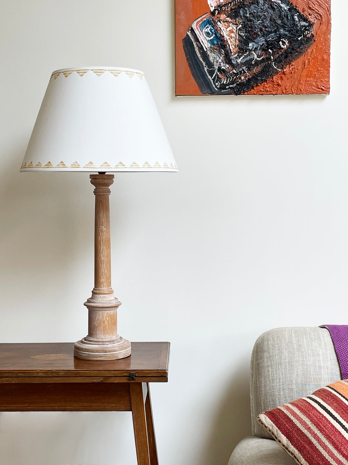 Styled sitting room scene with a cream empire lampshade featuring a yellow triangle border. The geometric lampshade sits on a carved wooden lamp base beside a neutral-toned sofa and bold artwork, blending modern design with cosy decor.