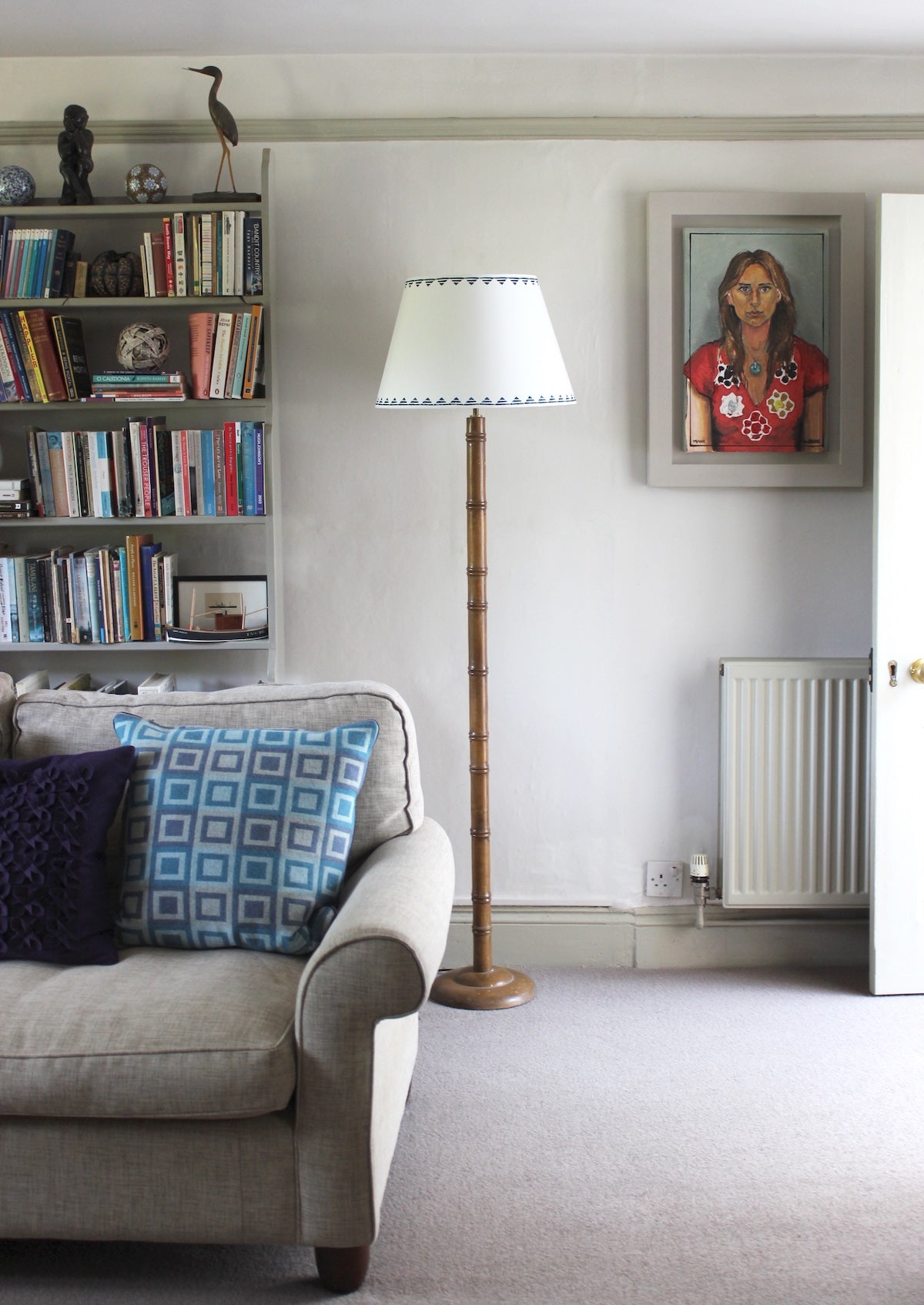 Living room interior full of sofa, bookshelf and portrait art, has a tall floor lamp fitted with a cream empire lampshade with a blue triangle pattern. This neutral lampshade with geometric detail adds a subtle colourful accent to the space.