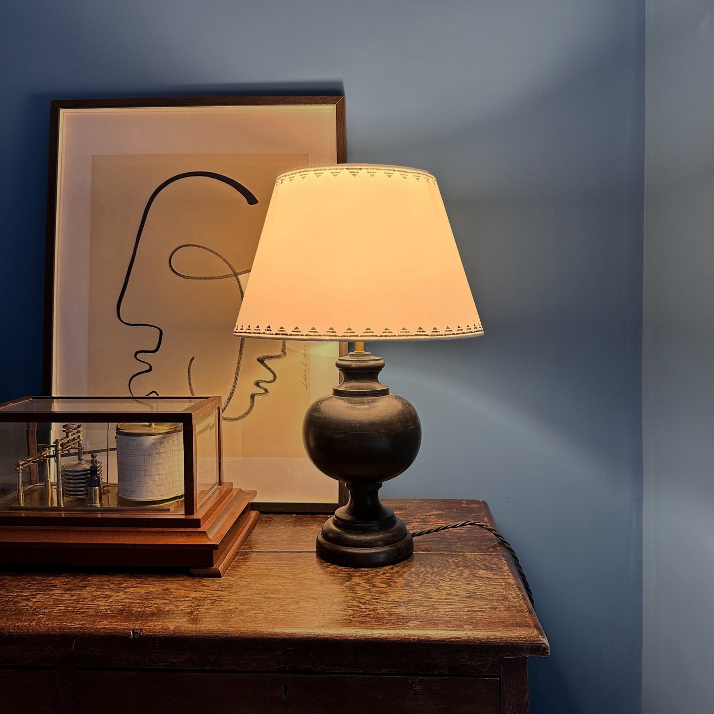 Romantic evening setting with a glowing cream empire lampshade featuring a lined triangle border in dark ink blue. The geometric lampshade casts a warm light, styled on a dark wooden lamp base against a deep blue wall.