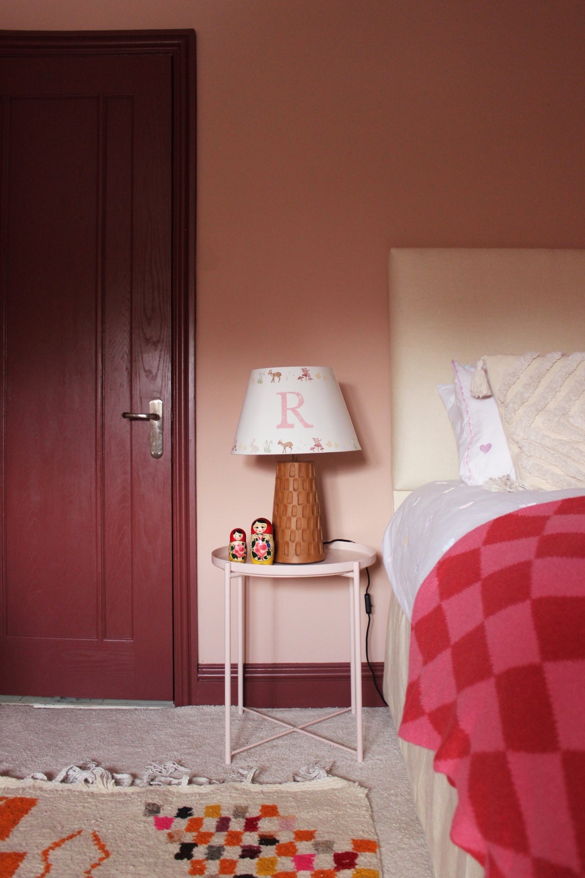 A cosy girls bedroom with a personalised children's lampshade featuring a woodland pattern and the letter "R" on a bedside table, alongside wooden nesting dolls. The decor is pink and red with a colourful rug and blanket.