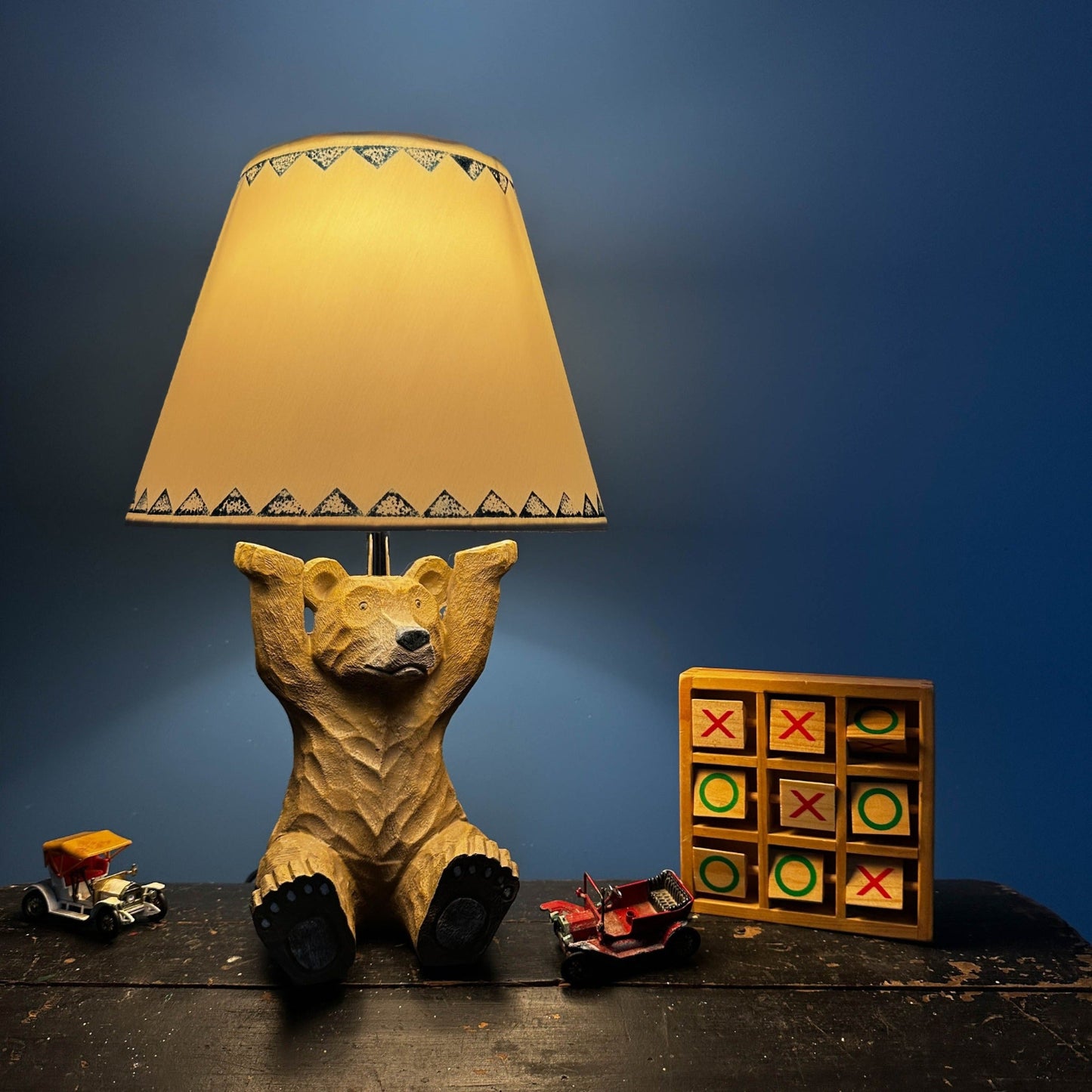 Warm glowing empire light shade with navy blue triangle border on a bear-shaped lamp base and antique wooden chest, a playful bold lampshade design suitable as a lampshade with triangle pattern for kids or cosy spaces. The table lamp is surrounded by vintage children's toys.