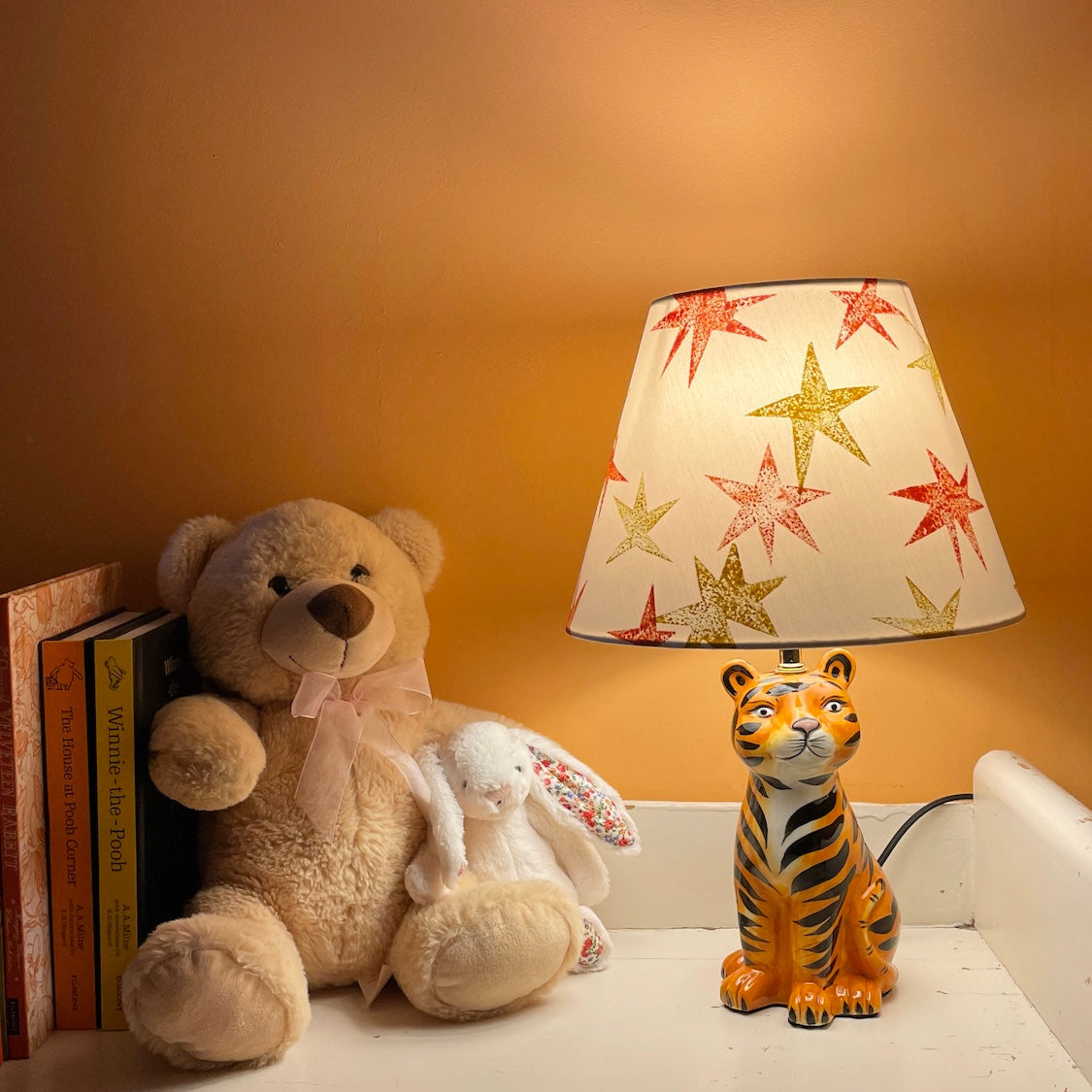 A dimly lit kids bedroom corner with a cream lampshade decorated with bright orange and yellow star patterns, styled in a kids bedroom on a tiger lamp base. A fun empire lampshade and kids bedroom decor piece that adds warmth and character.