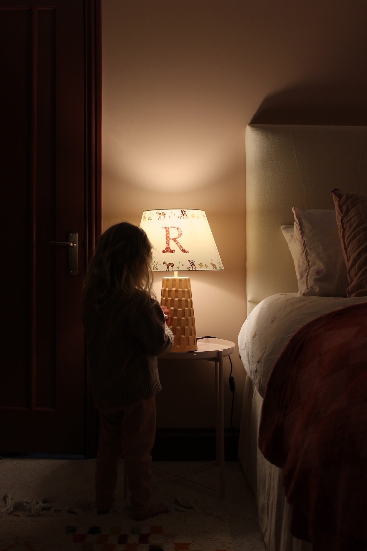A dimly lit bedroom with a glowing personalised lampshade featuring the letter "R" and a woodland pattern. A young girl stands beside it, reaching for the lamp.