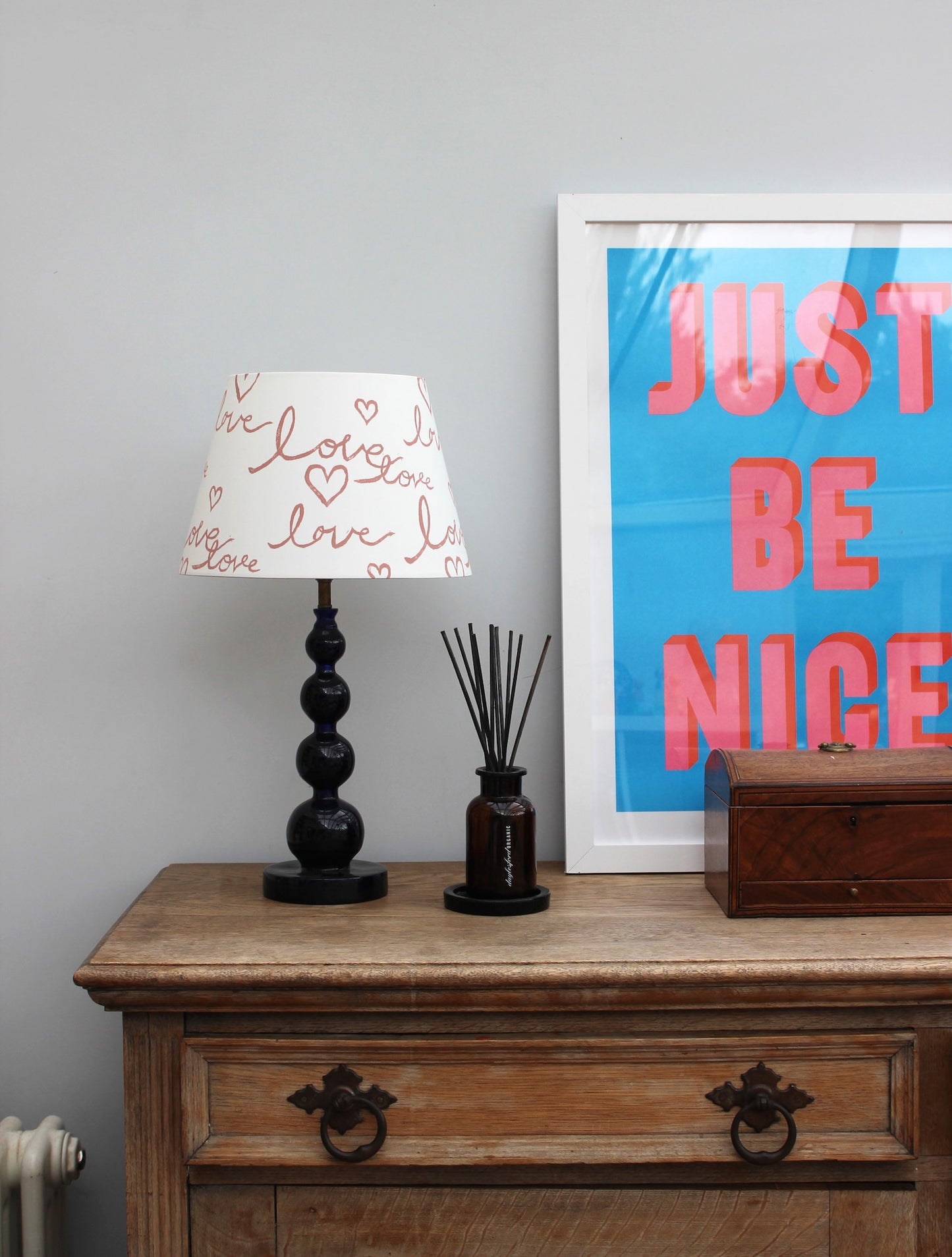 Studio Zef hand-painted lampshade with playful pink ‘Love’ lettering and hearts, styled on a blue table lamp against a modern interior with vintage wooden dresser and bold wall art.