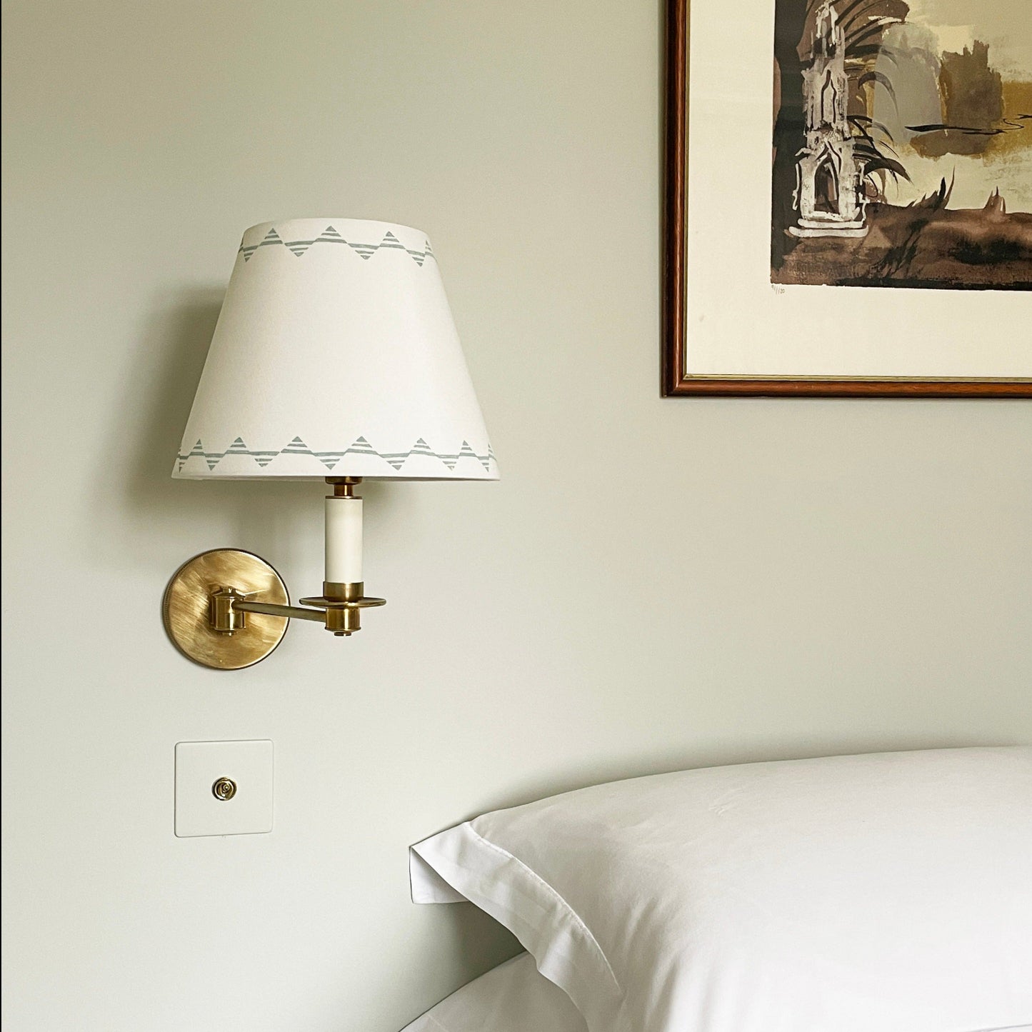 Neutral cream lampshade with triangle pattern on a brass wall-mounted bedside light, styled above a white bed with soft pillows.