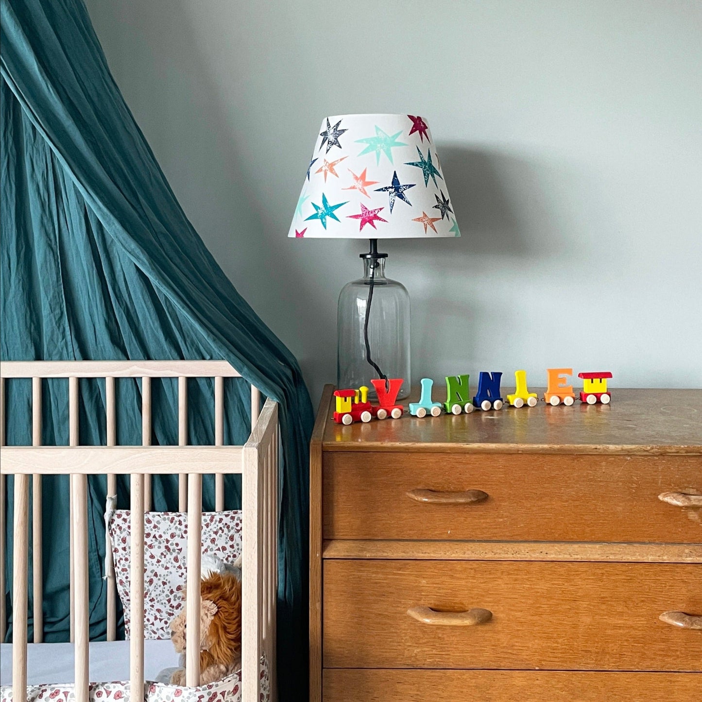 A colourful lampshade with multicoloured star prints, styled on a glass base place on a wooden chest of drawers next to a cot. This empire shade for table lamps is a cheerful lampshade with stars, perfect for kids bedroom decor.