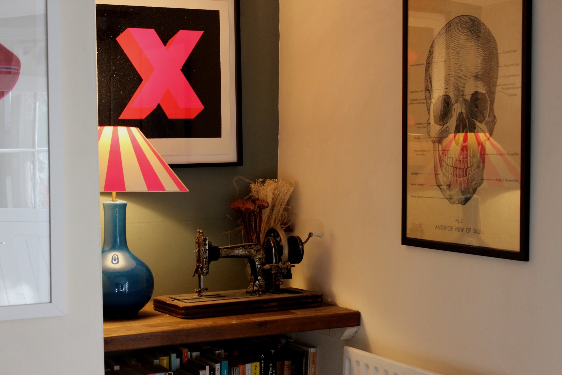 A cosy sitting room nook, with an eclectic mix of art on the walls including an antique looking anatomy of the skull and a neon pink X surrounded by black glitter. On the shelf in the nook is a blue lamp with a cone shaped lampshade which is decorated with neon pink stripes. Next to the lamp is a vintage singer sewing machine and vase of dried flowers.  