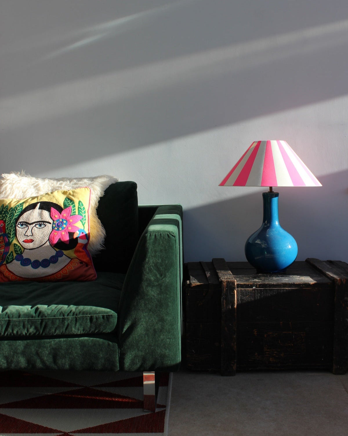 Sitting room corner with a large green velvet chair and antique chest as a side table. On the chest is a blue lamp with a cone shaped lampshade. The lampshade has bold and bright design of neon pink stripes.