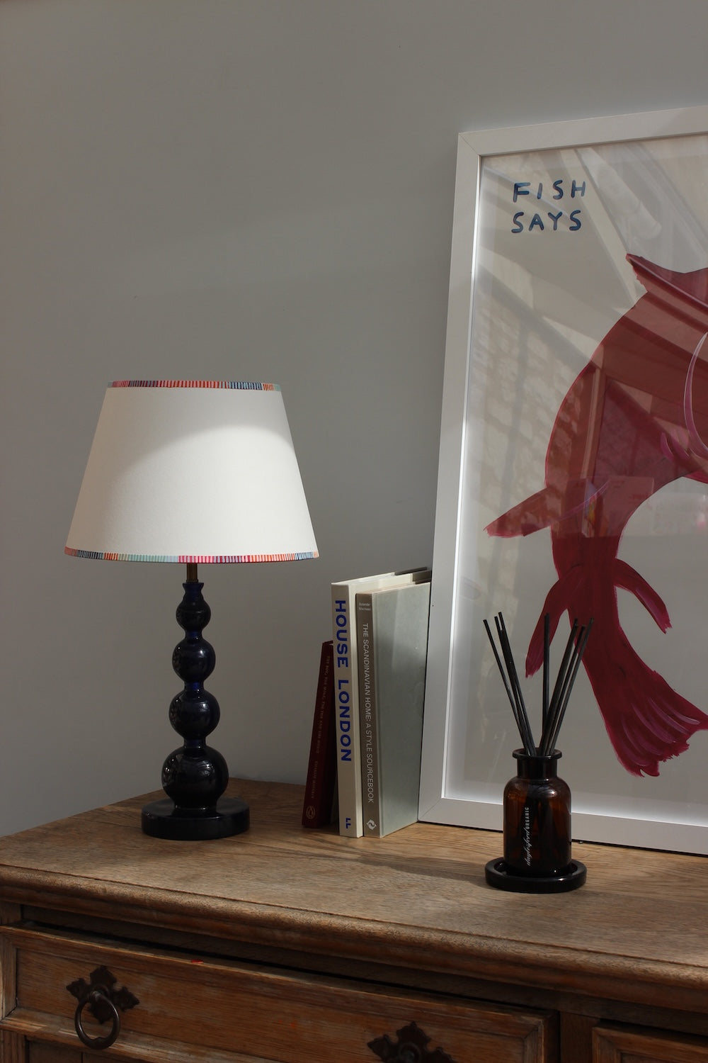Empire lampshade with bold, colourful stripe trim, styled on a dark blue table lamp with books and artwork. A striped lampshade that blends classic empire light shades with a modern colourful twist.