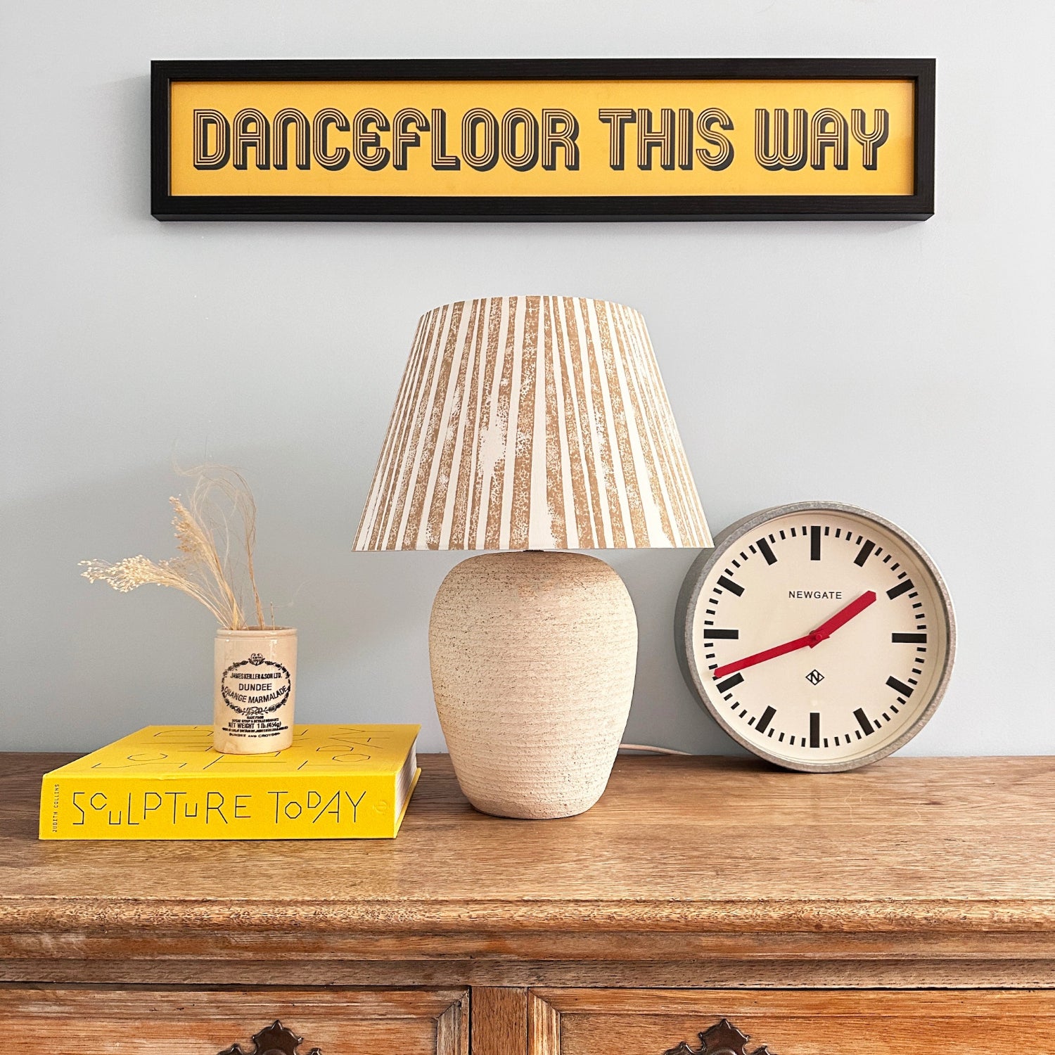 Bold stripe lampshade in warm neutral tones displayed on a rustic wooden sideboard, styled with a clock and books. A modern empire lampshade that combines striped lighting design with timeless elegance.