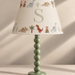 A children's table lamp with a pale green beaded stem and round base, paired with a cream-colored conical lampshade printed with colorful dinosaurs, cacti, campfires, and a large light green monogram letter 'S'.