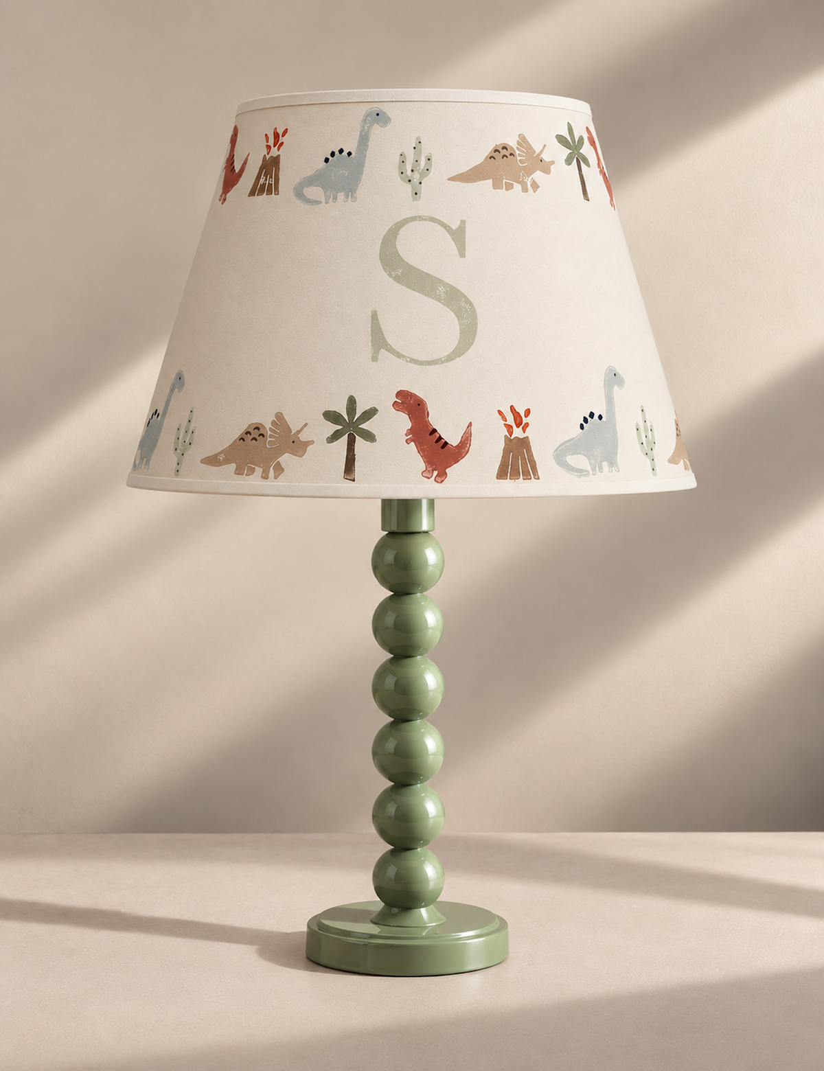 A children's table lamp with a pale green beaded stem and round base, paired with a cream-colored conical lampshade printed with colorful dinosaurs, cacti, campfires, and a large light green monogram letter 'S'.