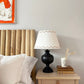Empire lampshade for table lamps in cream with geometric triangle design, styled on a wooden bedside table with books, black lamp base and white bedding. There is bight orange and white artwork above the bed.