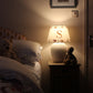 Glowing personalised bear lampshade with letter 'S', styled as kids lighting on a bedside table with teddy bears. A warm and neutral empire lampshade for kids bedroom décor. The rustic bedside table adored with a ceramic lamp and lampshade. There are two teddy bears, one on the bedside table next to the lamp, the other folded into the duvet. 