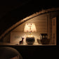 Children’s bedside table lamp with a personalised initial “T” lampshade, softly glowing in a cosy nursery setting. The handmade lampshade features small illustrated animal motifs around the rim, creating warm ambient lighting beside wooden toys and a dollhouse under a sloped ceiling.
