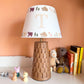 Personalised kids lampshade with grizzly bears and trees design, colourful empire lampshade with initial 'T' on a brown ceramic lamp base. A pretty lampshade for kids bedroom décor and new parent gifts. The lamp is in a kids bedroom chest of drawers next to stacked children's books, toys and soft toys.