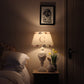 A dimly lit cosy children's bedroom with a rustic wooden nightstand featuring a personalised lampshade with the letter "C" and a countryside pattern. The lamps is on giving the room a soft ,warm glow. A plush goose, daffodils, and white bedding add charm.