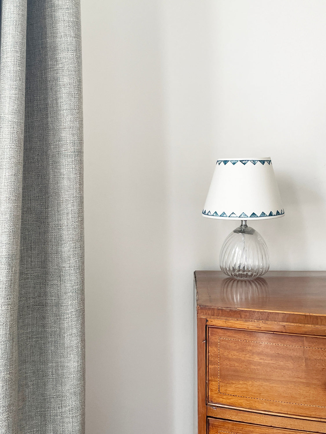 Small empire lampshade with geometric pattern, perfect for table lamps. This cream lampshade is placed on a wooden dresser with glass base, a bold lampshade with blue triangle border, perfect as a neutral lampshade in minimalist decor.