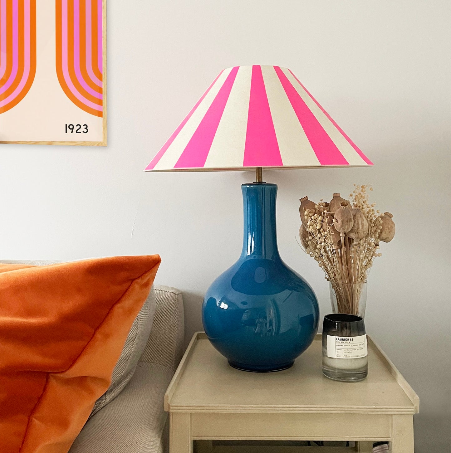 Living room scene featuring a vibrant pink and white striped lampshade atop a glossy blue ceramic lamp base on a beige side table. The table also holds a glass vase with dried flowers and a scented candle. An orange velvet cushion rests on a light grey sofa, and retro artwork is visible on the wall in the background