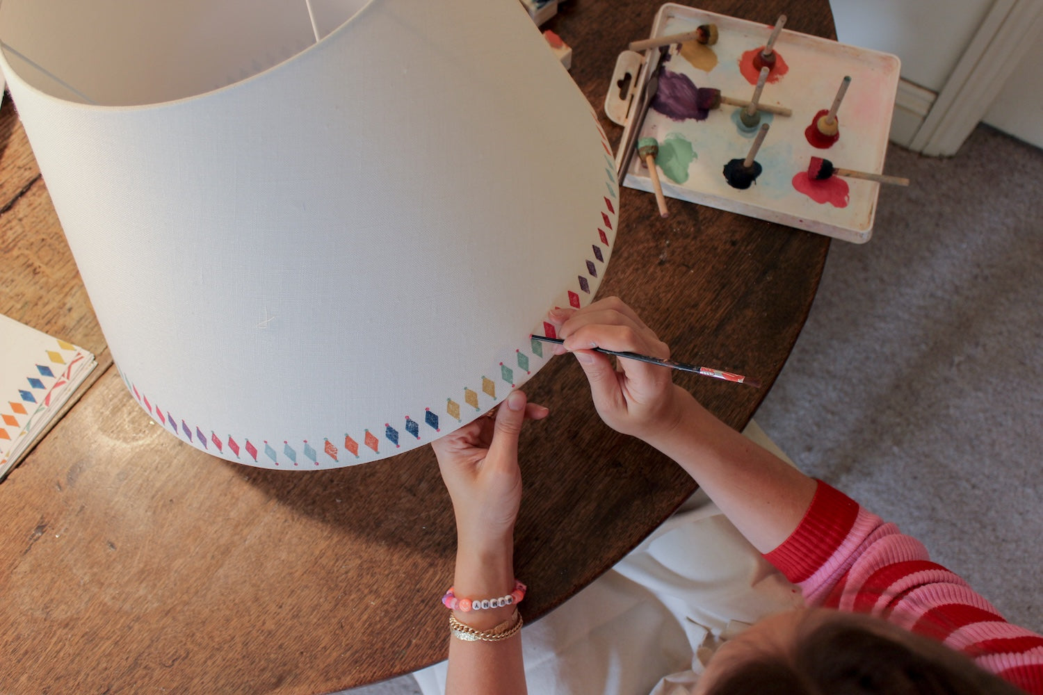 Grace, founder of Studio Zef, hand-printing a colourful empire lampshade for the Studio Zef x Kit Kemp collaboration. The pale grey lampshade features a bold, colourful geometric border with hand-painted diamond shapes, showcasing artisanal craftsmanship and unique lampshade design.