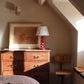 Cozy bedroom corner with warm natural light, featuring a wooden dresser, a red table lamp with a "love" pattern lampshade, a framed artwork, and a vintage wooden chair. A small reading nook with a colourful cushion is situated by the window.