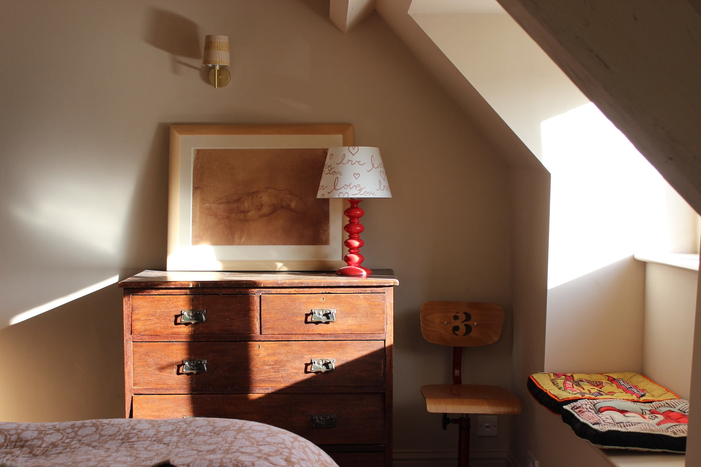 Cozy bedroom corner with warm natural light, featuring a wooden dresser, a red table lamp with a "love" pattern lampshade, a framed artwork, and a vintage wooden chair. A small reading nook with a colourful cushion is situated by the window.