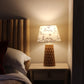 Cozy bedroom setting with a wooden headboard, plush pillows, and a bedside table featuring a warm glowing lamp. The empire-shaped lampshade is block-printed with the handwritten word "love" and heart motifs, adding a romantic and inviting touch to the space.
