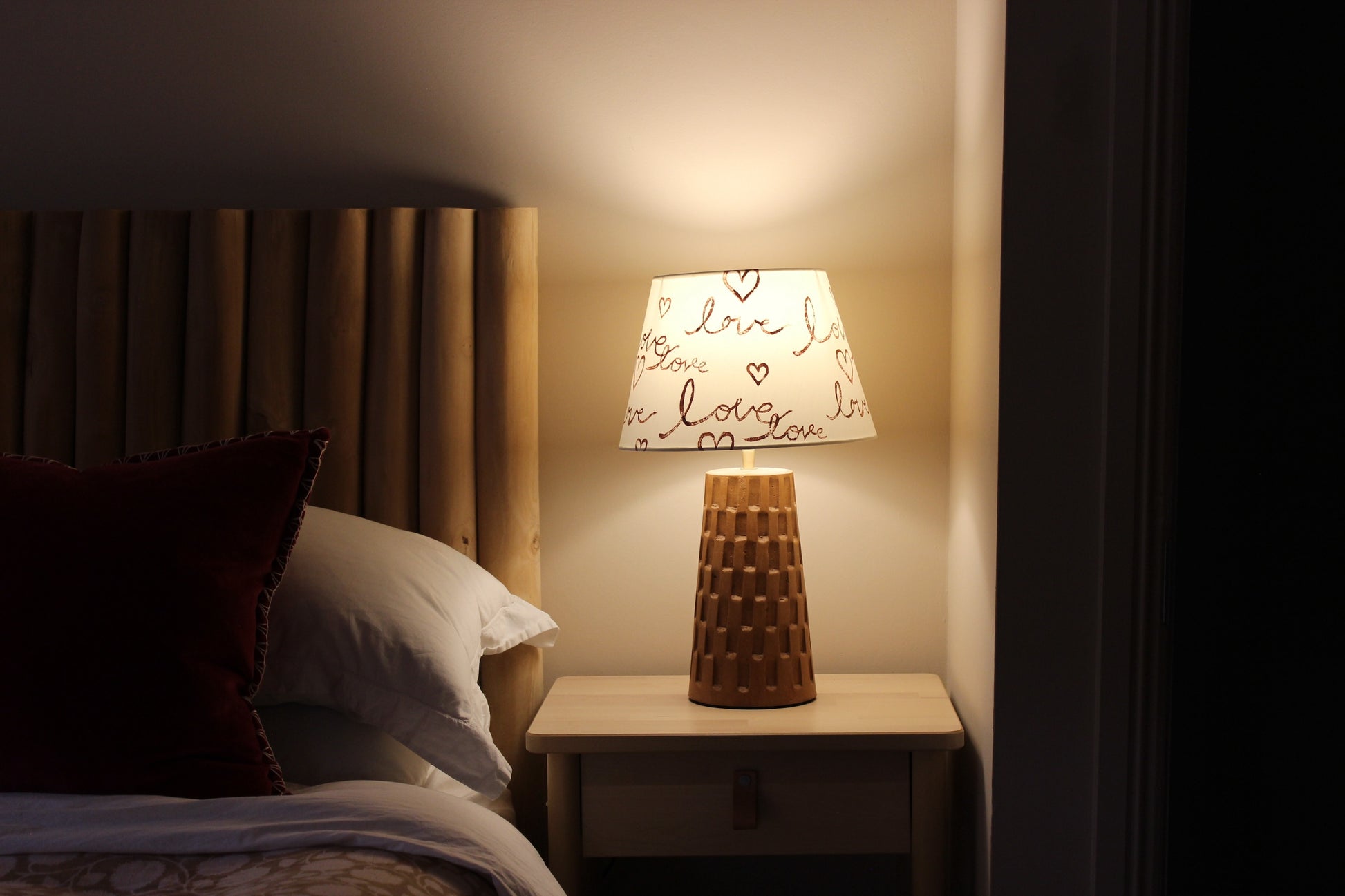 Cozy bedroom setting with a wooden headboard, plush pillows, and a bedside table featuring a warm glowing lamp. The empire-shaped lampshade is block-printed with the handwritten word "love" and heart motifs, adding a romantic and inviting touch to the space.