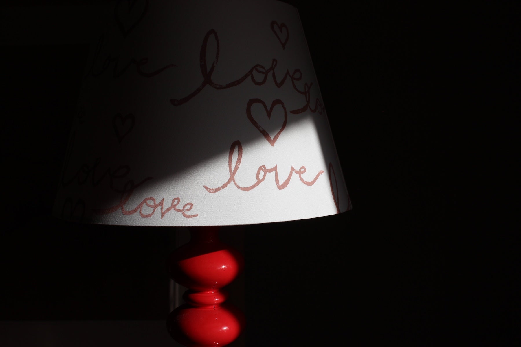 A close up image of an empire shaped lampshade with the hand written word 'Love' hand printed on to it with heart motifs. The lighting is natural but at stark contrast highlighting the word Love on the lampshade. 