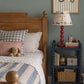 A styled children's bedroom corner featuring a wooden bed with blue and white striped bedding, a navy blue nightstand with a red lamp and books, and a framed tractor print on a muted blue-gray wall.