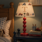 A personalized children's table lamp with a red stacked base and a cream fabric shade printed with farm tractors, a barn, and a large letter 'T', shown lit on a dark blue nightstand in a cozy bedroom setting.