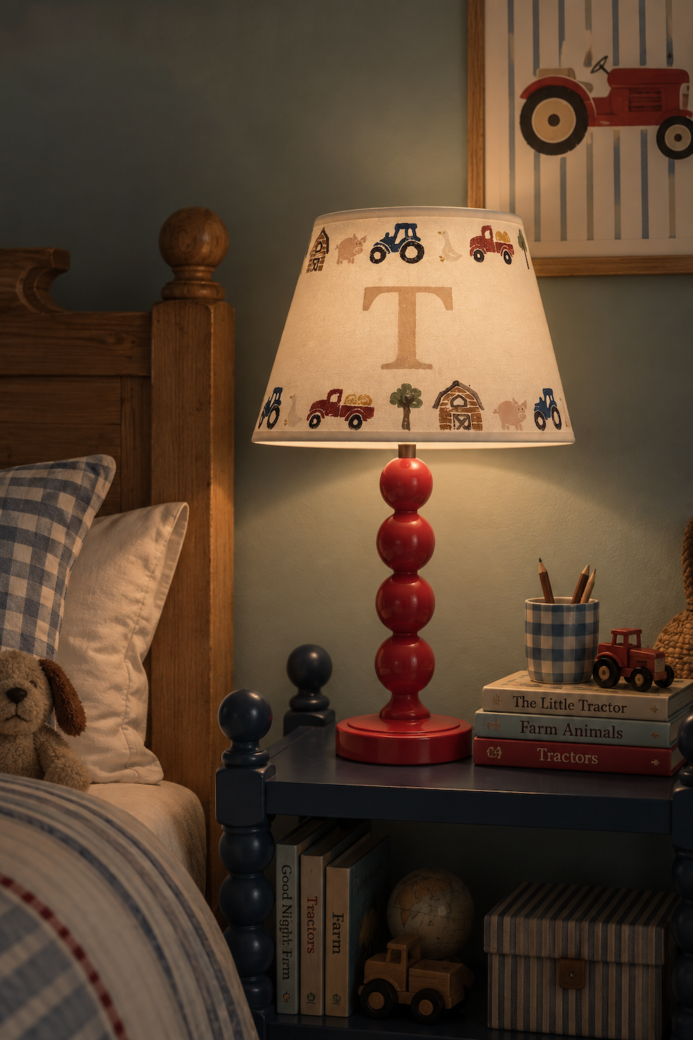 A personalized children's table lamp with a red stacked base and a cream fabric shade printed with farm tractors, a barn, and a large letter 'T', shown lit on a dark blue nightstand in a cozy bedroom setting.