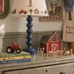 A styled nursery or children's room scene showing a blue turned wood table lamp with a cream fabric shade decorated with colorful farm vehicles and a large letter 'T'. The lamp sits on a distressed sage green dresser alongside toy books, a red barn figurine, small animal toys, and framed wall art.