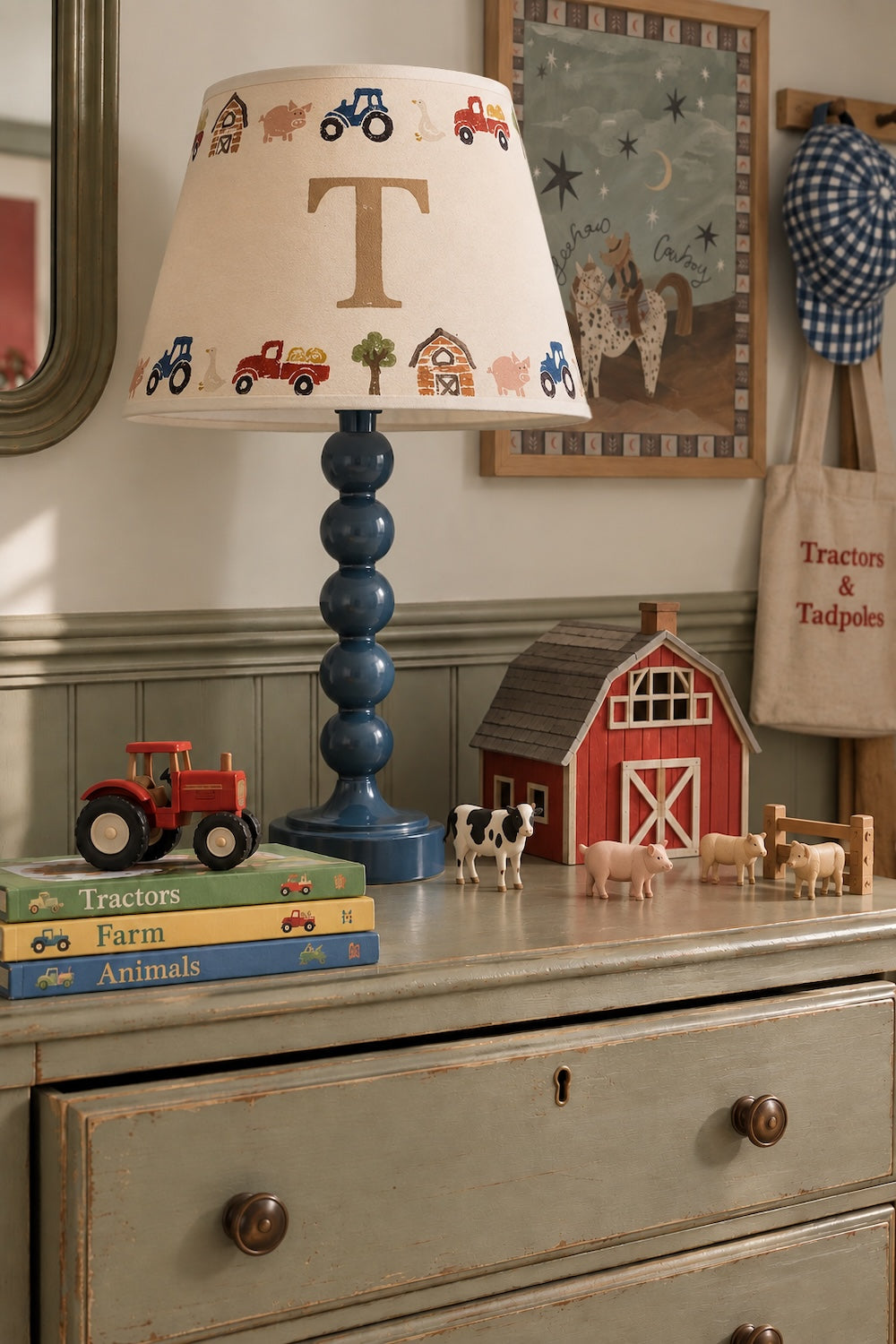 A styled nursery or children's room scene showing a blue turned wood table lamp with a cream fabric shade decorated with colorful farm vehicles and a large letter 'T'. The lamp sits on a distressed sage green dresser alongside toy books, a red barn figurine, small animal toys, and framed wall art.