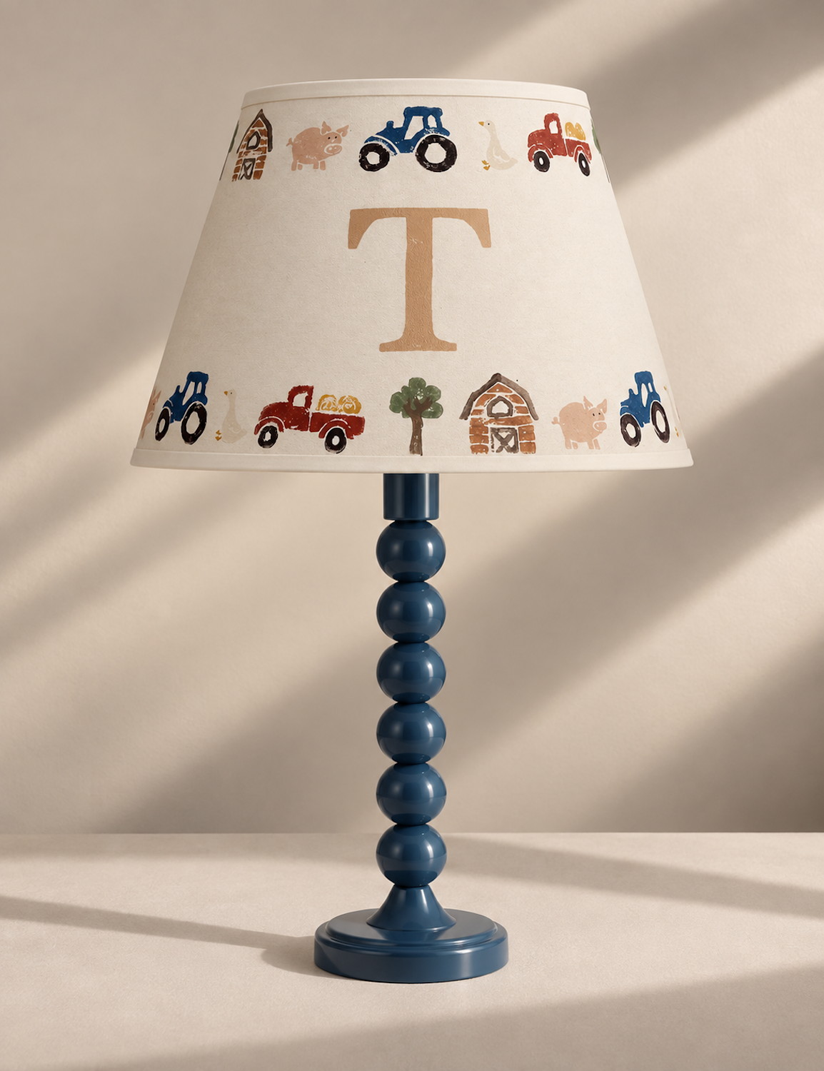 A personalized farm-themed table lamp with a blue beaded stem and a cream fabric shade decorated with colorful tractors, a pig, a goose, and a large gold initial 'T'.