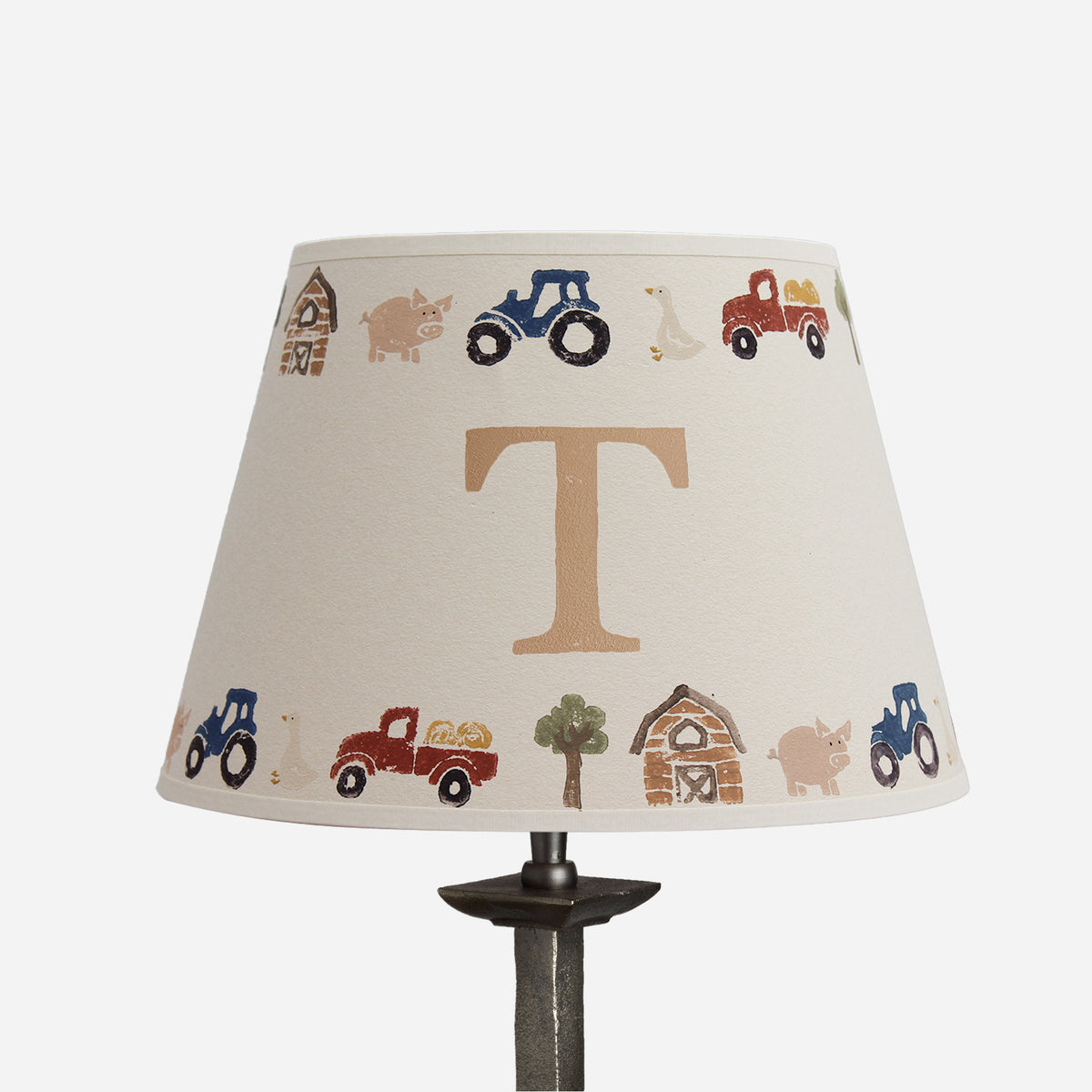 A table lamp shade with a cream fabric background printed with a circular farm-themed pattern of tractors, a barn, a pig, a tree, and a large tan letter T in the center, shown on a dark lamp stand against a light gray background.