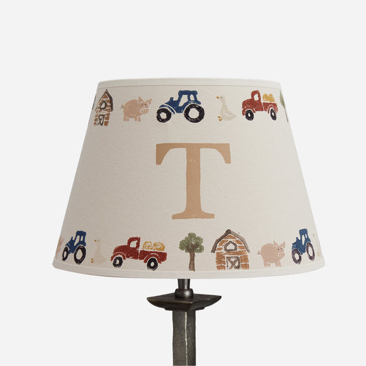 A table lamp shade with a cream fabric background printed with a circular farm-themed pattern of tractors, a barn, a pig, a tree, and a large tan letter T in the center, shown on a dark lamp stand against a light gray background.