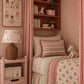 A cozy children's bedroom featuring a single bed dressed in pink and green patterned bedding, a matching canopy with scalloped trim, a pink nightstand with a wicker lamp, and a built-in wooden shelf with books and small decor items.